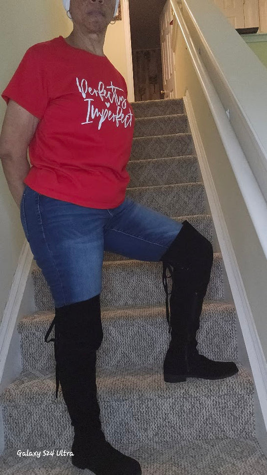 Person wearing a red shirt with white text, blue jeans, and black over the knee boots on a staircase.